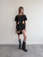 Person wearing a black outfit with ripped elements and black boots against a plain background