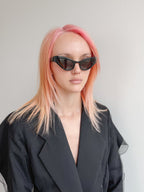 Person with pink hair wearing black sunglasses and a black coat against a light gray background