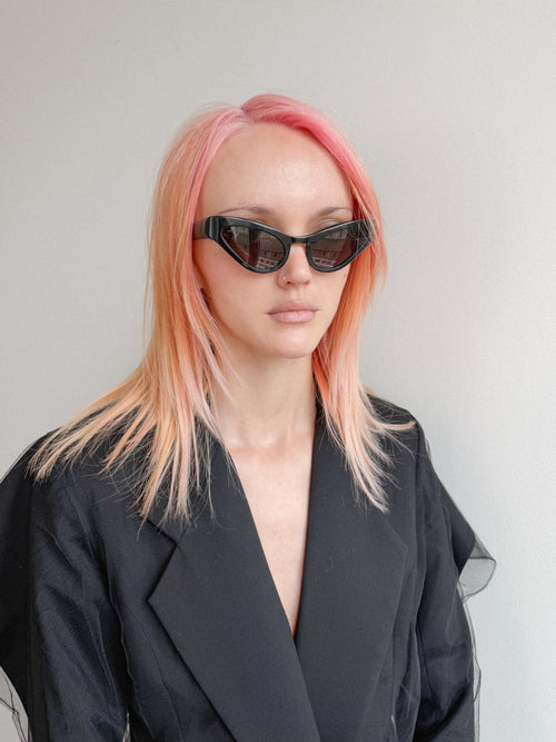 Person with pink hair wearing black sunglasses and a black coat against a light gray background