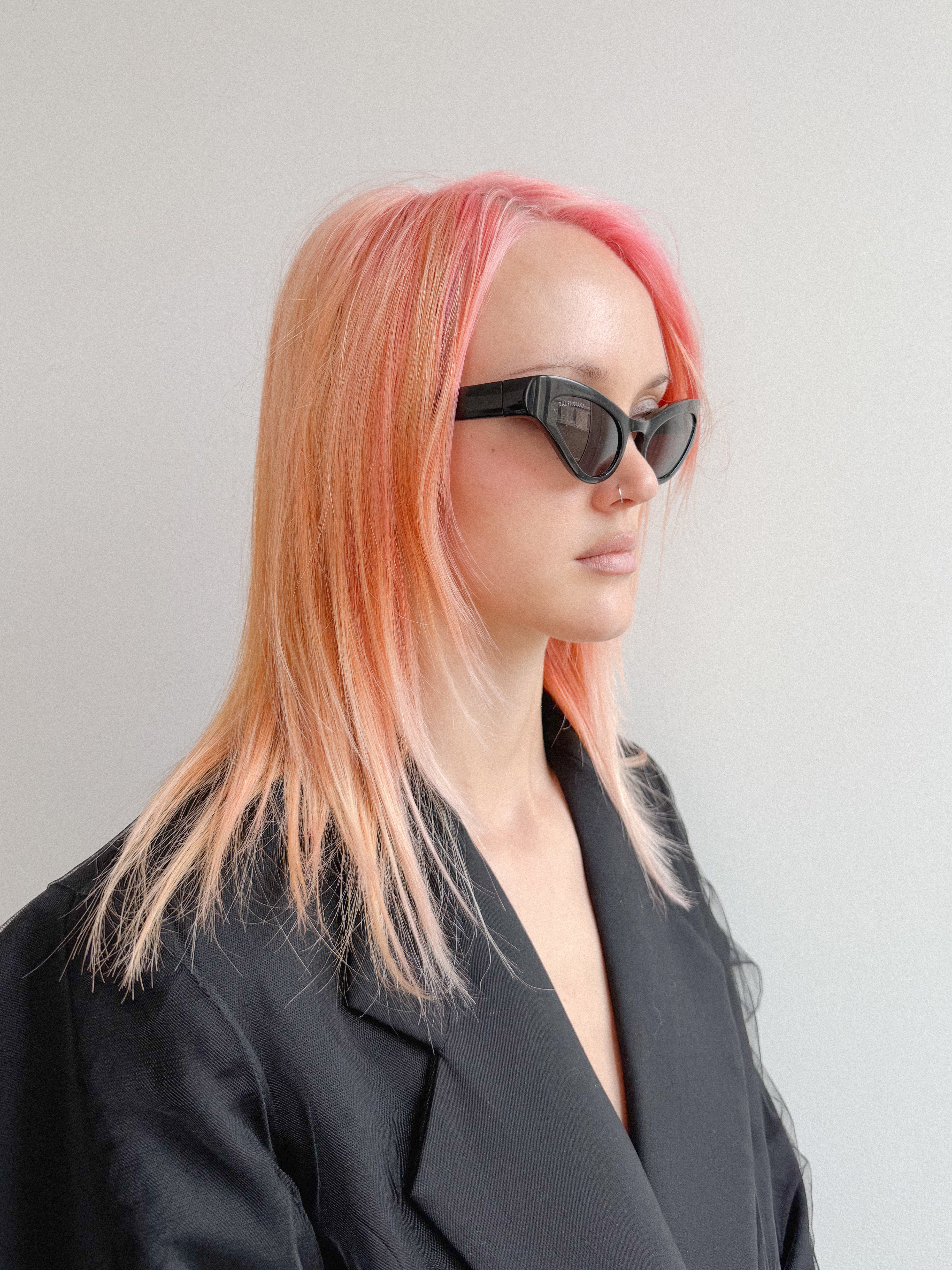 Person with pink hair wearing black sunglasses and a black outfit against a gray background