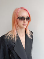 Person with pink hair wearing sunglasses and a black outfit against a plain background