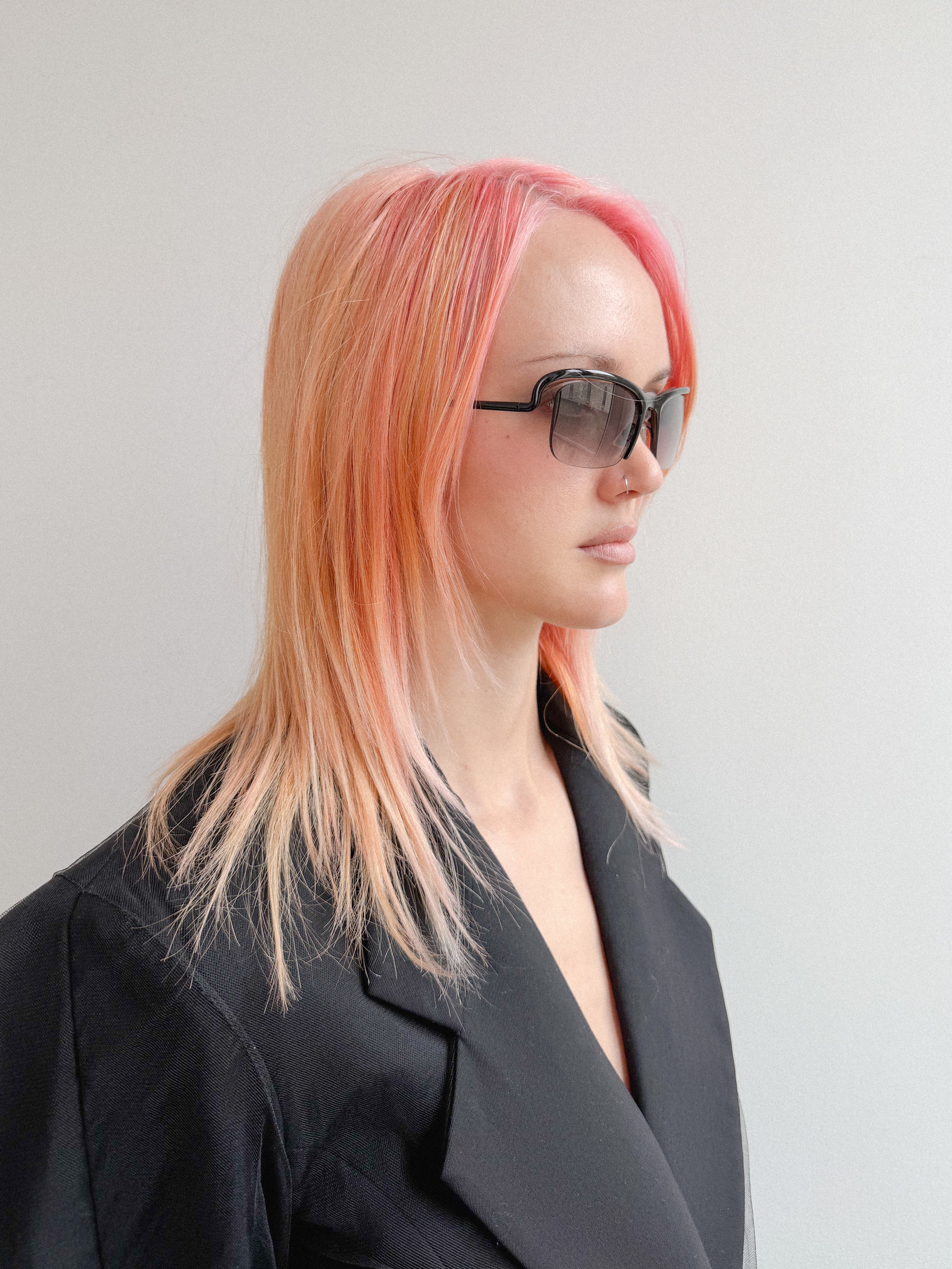 Person with pink hair wearing sunglasses and a black outfit against a plain background
