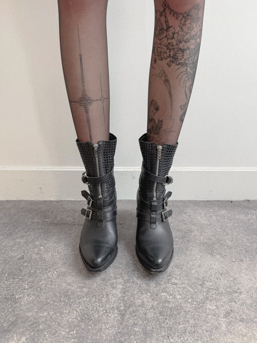 Black leather boots with buckles worn with sheer black tights against a neutral background