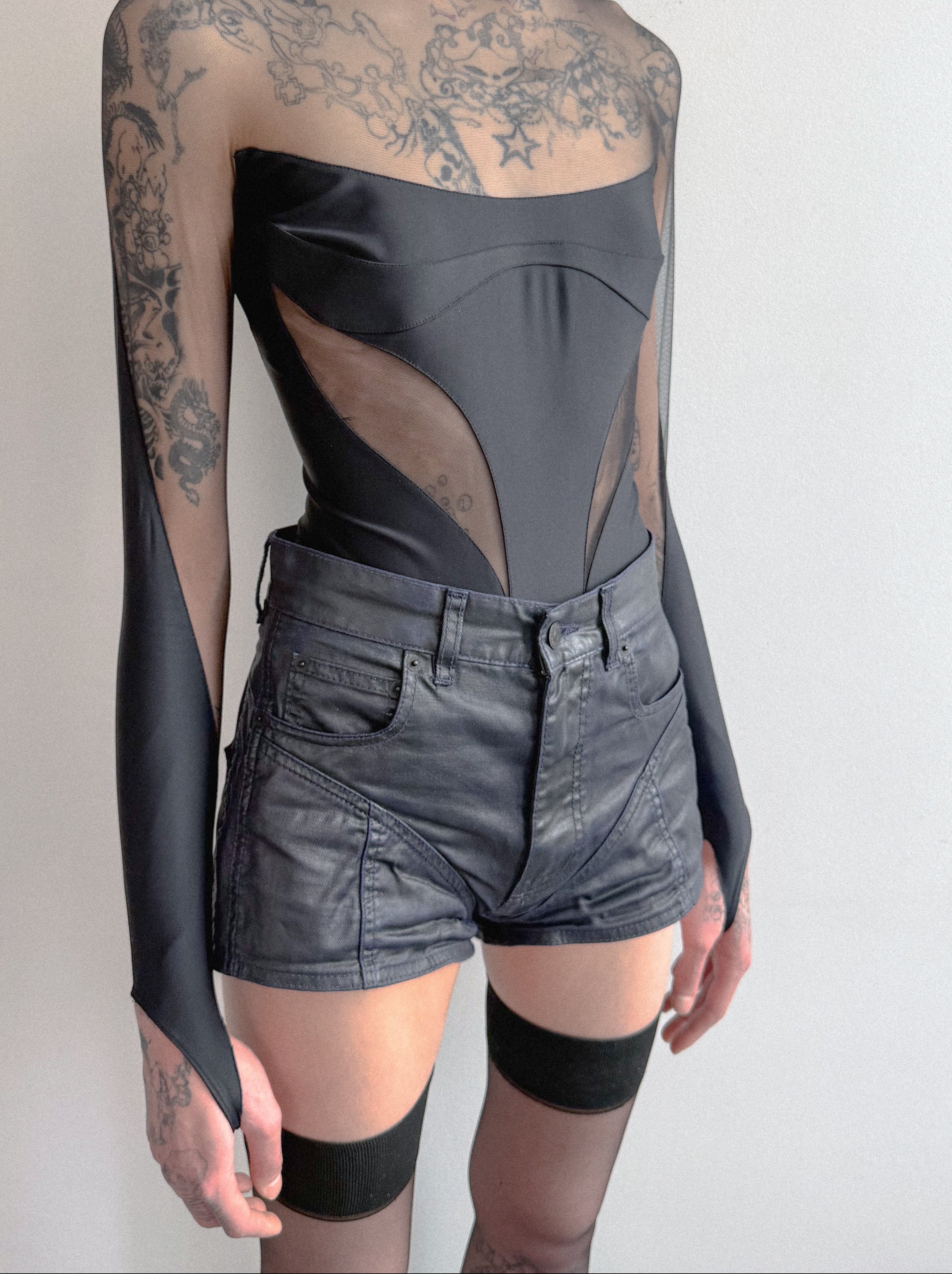 Person wearing a sheer long-sleeve top with floral patterns and black high-waisted shorts on a plain background.