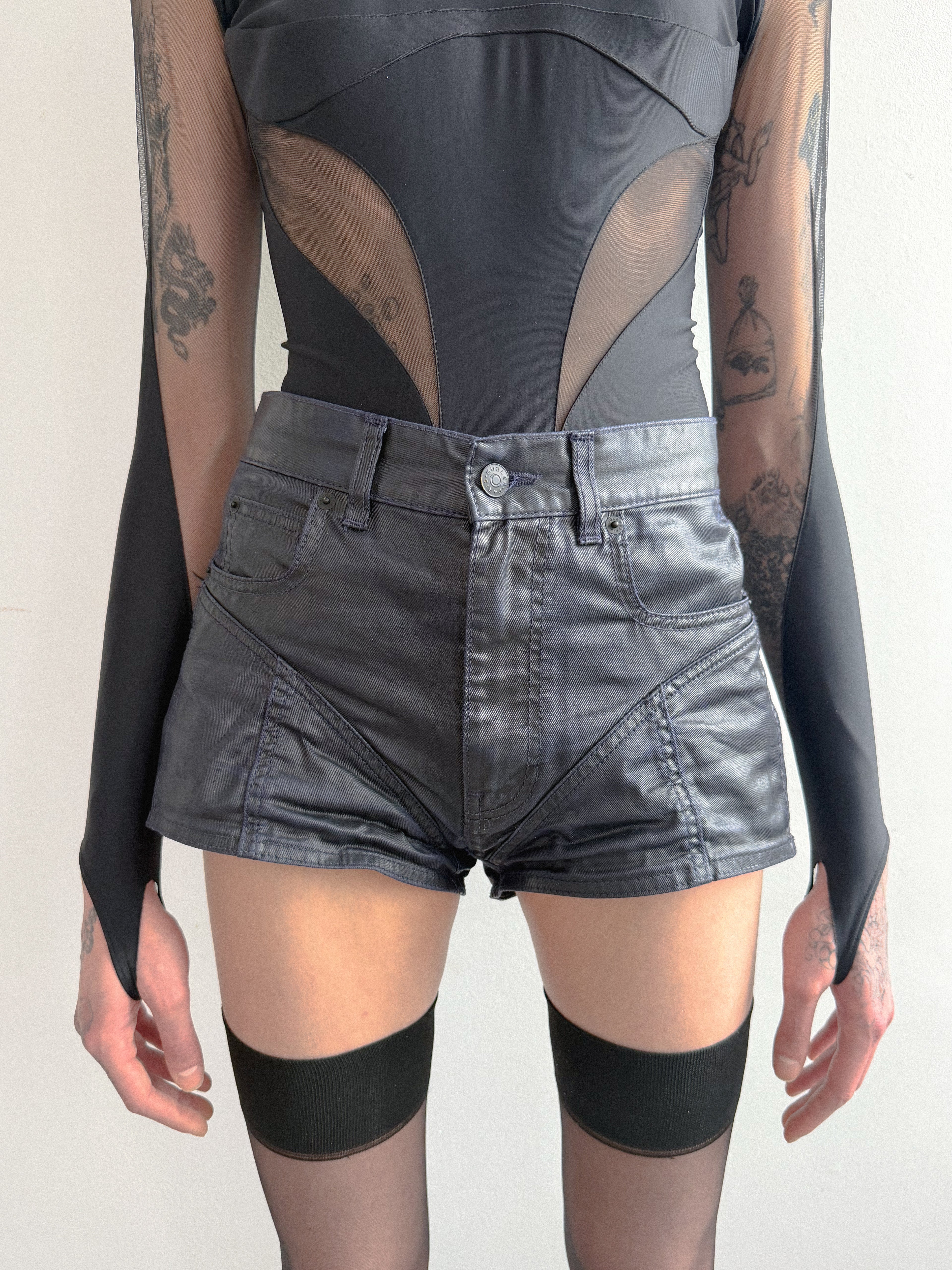 Person wearing a black bodysuit with sheer panels and high-waisted black shorts on a plain background