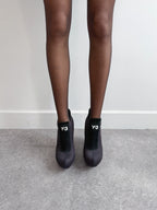 Black high-heeled shoes with 'Y-3' logo worn by a person on a gray floor.