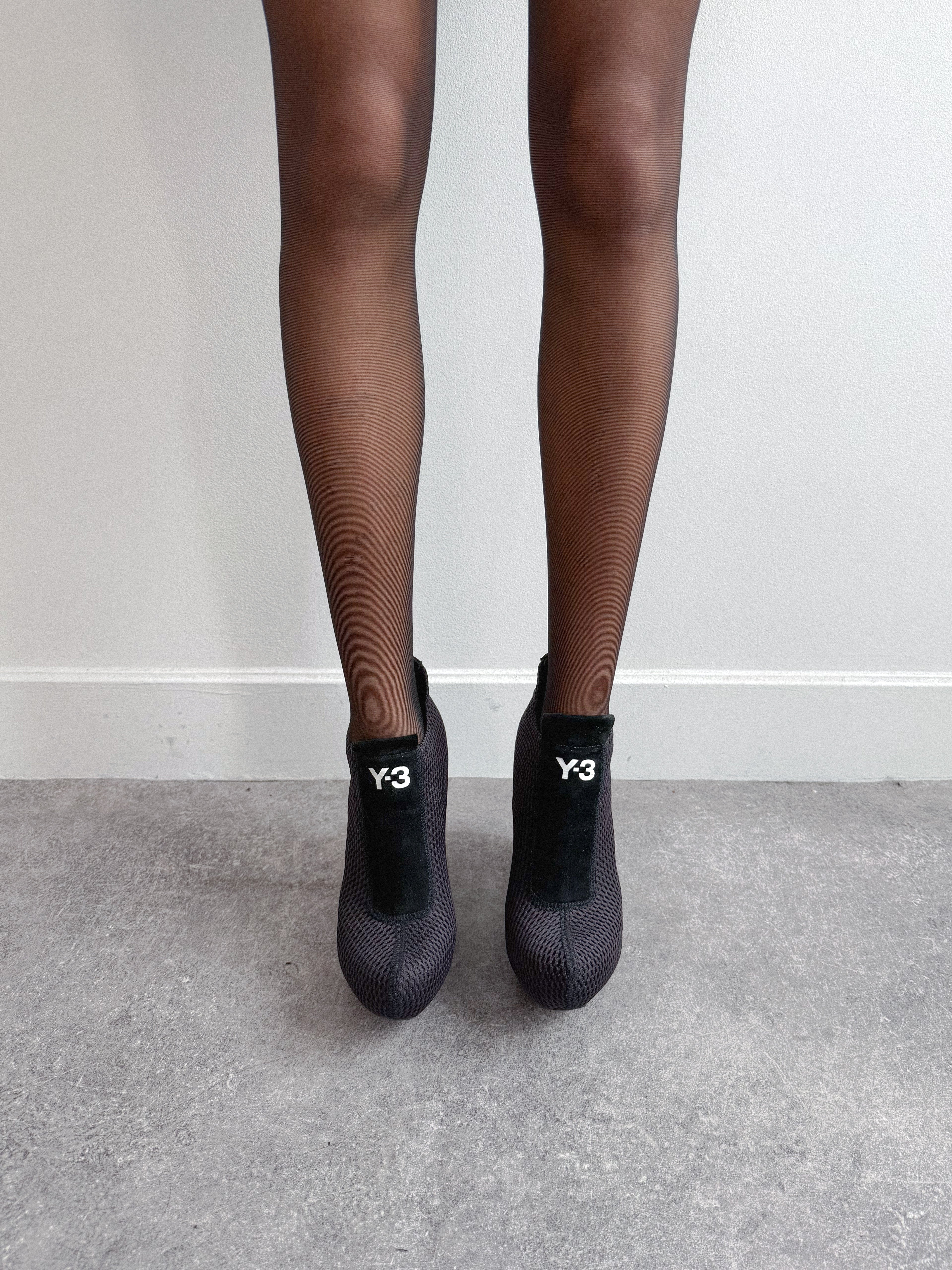 Black high-heeled shoes with 'Y-3' logo worn by a person on a gray floor.