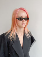 Person with pink hair wearing sunglasses and a black coat against a gray background