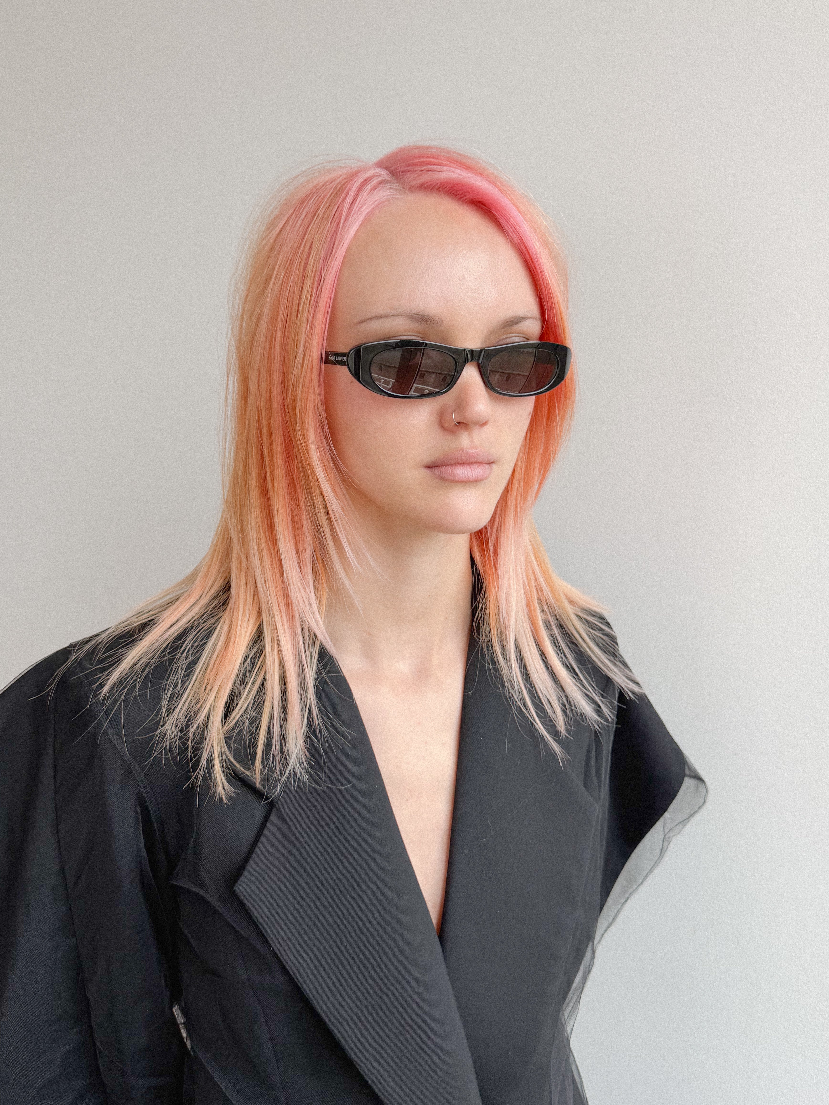 Person with pink hair wearing sunglasses and a black coat against a gray background