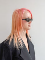 Person with pink hair wearing sunglasses and a black outfit against a gray background